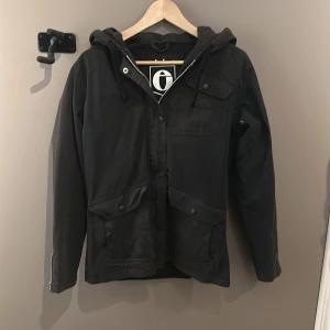 Svart jacka från Elvine - Snygg svart jacka från Elvine med huva och dragkedja. Jackan har praktiska fickor och knappar framtill. Perfekt för kyligare dagar.