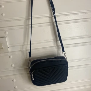 Svart quiltad axelväska med dragkedja - Snygg svart axelväska med quiltad design och dubbla dragkedjor. Perfekt för att bära dina nödvändigheter med stil. Justerbar axelrem för bekväm passform.