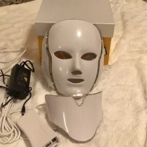 LED-ljusbehandlingsmask - Säljer en LED-ljusbehandlingsmask med ansikts- och halsdel. Masken är vit och genomskinlig med inbyggda LED-lampor. Den kommer med en fjärrkontroll för att justera inställningar och en strömadapter. Perfekt för hudvård och avkoppling hemma.