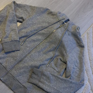 Grå hoodie med dragkedja - Säljer en bekväm grå hoodie med dragkedja framtill. Tröjan har långa ärmar och ribbade muddar vid ärmslut och nederkant. Perfekt för en avslappnad stil.