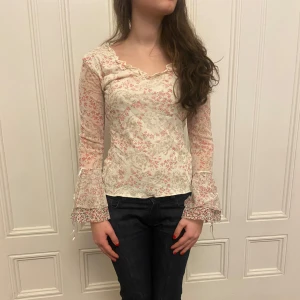Blommig långärmad blus - Säljer en söt blommig blus med lång ärm och volangdetaljer. Blusen har en v-ringad hals och är i en ljus beige färg med rosa blommönster. 