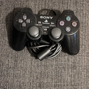 Sony PlayStation 2 med handkontroll - Säljer en klassisk Sony PlayStation 2 i svart med tillhörande handkontroll. Perfekt för retrospel och samlare. Konsolen har den ikoniska PS2-loggan i blått och lila på ovansidan. Handkontrollen har de klassiska PlayStation-knapparna och en bekväm design.