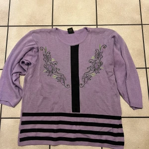Lila tröja med broderi storlek M/L - Säljer en lila tröja med vackert broderi framtill och svarta ränder längst ner. Perfekt för en stilren look.