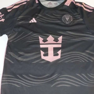 Svart fotbollströja från Adidas - Snygg svart fotbollströja från Adidas med rosa detaljer och klubbmärke på bröstet. Tröjan har korta ärmar och ett unikt vågmönster. Perfekt för fotbollsfans! Storleken är S och skicken är riktigt bra