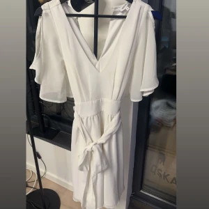 Vit klänning med knytband - Elegant vit klänning från bubbleroom med v-ringning och knytband i midjan. Den har kortärmade ärmar och en luftig passform, perfekt för sommarens festligheter eller studentfirande.