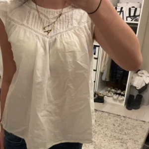 Vit blus med spetsdetaljer - Säljer en söt vit blus med spetsdetaljer och rund hals. Blusen har en luftig passform och är perfekt för varmare dagar. Den är ärmlös och har en fin struktur i tyget.