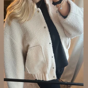 Beige bomberjacka  - Mysig beige bomberjacka i boucle material med knappar framtill och två stora fickor. Perfekt för att hålla sig varm och stilren under kyligare dagar. Jackan har en croppad passform och långa ärmar.