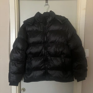 Svart pufferjacka - Snygg svart pufferjacka med dragkedja och huva. Perfekt för kyliga dagar med sin vadderade design. Jackan har långa ärmar och en glansig yta som ger en modern look, jackan har avtagbar huva!