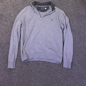 Grå tröja med dragkedja från Ralph Lauren - Säljer en stilren grå tröja från Ralph Lauren med halv dragkedja och broderad logga på bröstet. Perfekt för en avslappnad look. Tröjan har långa ärmar och ribbade muddar.