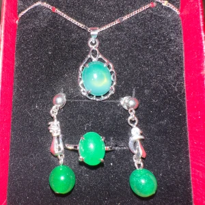 Smyckeset med gröna stenar - Elegant smyckeset med halsband, örhängen och ring. Varje del har en vacker grön sten infattad i silverfärgad metall. Halsbandet har en droppformad hängsmycke, örhängena är dinglande och ringen har en stor oval sten.