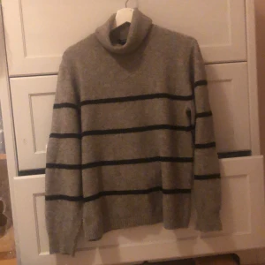 Grå randig stickad polotröja från Lindex - Säljer en stilren grå polotröja från Lindex med svarta ränder. Tröjan har långa ärmar och en bekväm passform, perfekt för kyligare dagar. Den är tillverkad i ett mjukt material som känns skönt mot huden.
