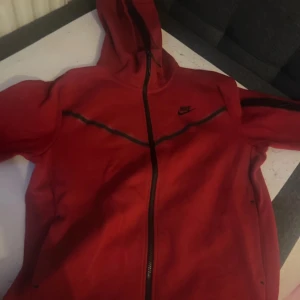 Röd hoodie från Nike - Snygg röd hoodie från Nike med dragkedja och huva. Perfekt för en sportig look. Svart Nike-logga på bröstet och svarta detaljer längs ärmarna.