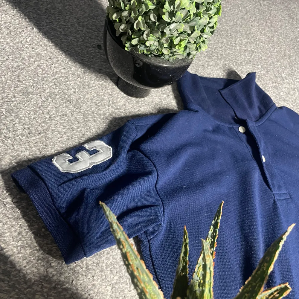 Säljer en snygg mörkblå pikétröja från Ralph Lauren med klassisk krage och knappar. Tröjan har en stor vit logotyp på bröstet och en siffra på ärmen. Perfekt för en stilren look!. Neuletakit & Villapaidat.