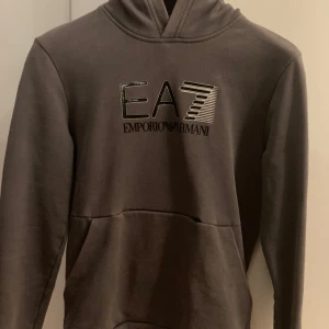 Grå hoodie från Emporio Armani - Säljer en stilren grå hoodie från Emporio Armani med EA7-logga på bröstet. Tröjan har en klassisk design med huva och en stor ficka framtill. Perfekt för en avslappnad look.