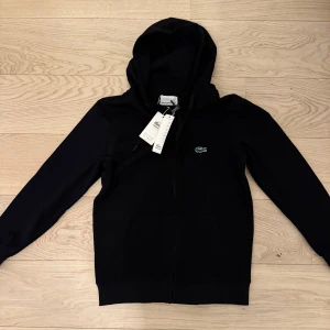 Lacoste Zip Up Hoddie - Jag säljer den här sprillans nya lacoste full zip. Jag säljer den pga storleken. Helt ny, inga defekter, 10/10 skick. Den passar XS men kan också passa S vid en tajt fit. Vid fler frågor eller bilder är det bara o höra av sig.