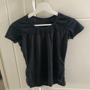 Svart träningstopp från Newbody - Säljer en svart träningstopp från Newbody. Den är kortärmad och har ett ventilerande meshmaterial på axlarna och ryggen, perfekt för träning. Toppen är i bra skick och har en normal passform.