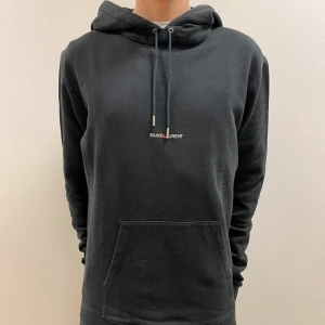 Svart hoodie från Saint Laurent - Säljer en stilren svart hoodie från Saint Laurent. Den har en klassisk design med en justerbar huva. Perfekt för en avslappnad och trendig look. Passar till alla tillfällen! modellen på bilden är 185cm