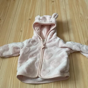 Rosa fleecejacka med huva och öron - Supergullig rosa fleecejacka för barn med huva som har små öron. Jackan har ett hjärtmönster och dragkedja framtill. Perfekt för att hålla sig varm och mysig under kyliga dagar!