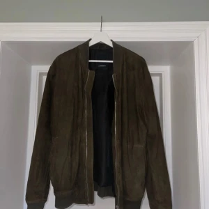 Jacka i mocka från J.Lindberg - Fin grön mockajacka från J.Lindberg i bomberjacka modell. Storlek L. Liten fläck vid kragen. Därav priset. Se sista bilden 