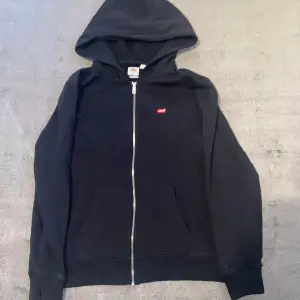 Säljer en klassisk svart hoodie från Levi's i storlek S. Den har en dragkedja framtill och en liten röd Levi's-logga på bröstet. Perfekt för en avslappnad stil och passar till det mesta. Superbekväm och stilren!