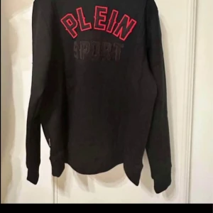 Svart hoodie från Plein Sport - Snygg svart hoodie från Plein Sport med dragkedja och röd text på ryggen. Perfekt för en sportig look. Den har långa ärmar och en stilren design med märkets logga på bröstet.