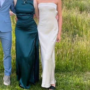 vit/Biege axelbandslös långklänning - Elegant vit axelbandslös långklänning med en stilren design. Klänningen har en smickrande passform och en öppen ryggdetalj som ger en extra touch av glamour. Perfekt för speciella tillfällen.