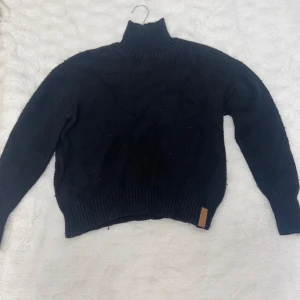 Svart stickad polotröja - Säljer en stilren svart stickad polotröja med ribbade muddar och en hög krage. Perfekt för kyligare dagar och ger en klassisk look. 