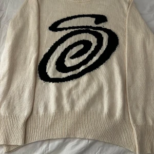Beige stickad tröja från Stüssy - Säljer en beige stickad tröja från Stüssy med ett stort svart spiraltryck på framsidan. Tröjan har en rund halsringning och långa ärmar. Perfekt för en stilren och avslappnad look.