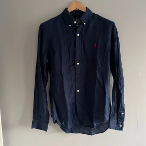 Mörkblå skjorta från Ralph Lauren - Snygg mörkblå skjorta från Ralph Lauren i slim fit. Skjortan har långa ärmar och knappar framtill samt en broderad röd logotyp på bröstet. Perfekt för en stilren look.