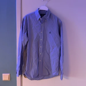 Blå rutig skjorta från Ralph Lauren - Snygg blå rutig skjorta från Ralph Lauren i storlek M. Skjortan har långa ärmar och klassiska knappar framtill. Den är broderad med den ikoniska logotypen på bröstet. Perfekt för en stilren look.