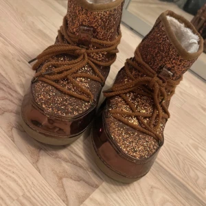 Bruna glittriga boots med snörning - Snygga bruna boots med glittrig yta och snörning. De har en varm, fodrad insida som passar perfekt för kallare dagar. Skorna har en blank finish och är i storlek 41 men passar troligtvis 40-42!