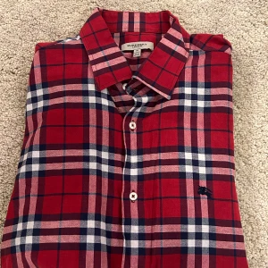 Äkta, Rutig röd skjorta från Burberry - Snygg röd rutig skjorta från Burberry med klassiskt mönster och broderad logga. Skjortan har långa ärmar och knappar framtill. Perfekt för en stilren look.