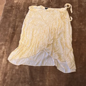 Gul omlottkjol från Lindex - Säljer en söt gul omlottkjol från Lindex med ett blommigt mönster. Kjolen har en knytning i midjan och en fin volangdetalj som ger en lekfull look. Perfekt för varma dagar!