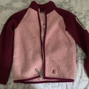 Rosa teddyjacka från 157 Funk - Mysig rosa teddyjacka från 157 Funk med vinröda ärmar och detaljer. Jackan har en dragkedja framtill och är perfekt för kyliga dagar. Den är långärmad och har en skön, fluffig textur som håller dig varm och bekväm.