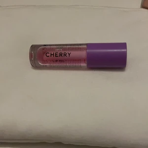 Cherry Lip Oil från H&M - Säljer ett Cherry Lip Oil från H&M med en söt körsbärsdoft. Den har en lila kork och en genomskinlig flaska som visar den rosa oljan inuti. Perfekt för att ge läpparna en glansig finish och extra fukt. Helt ny och har inte använts.