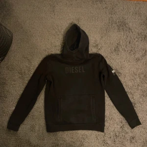 Svart hoodie från Diesel - Säljer en stilren svart hoodie från Diesel med logga på bröstet och en patch på ärmen. Perfekt för en casual look med sin bekväma passform och klassiska design. Huvan ger extra mysfaktor och den har en praktisk magficka framtill.