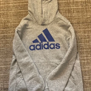 Grå hoodie från Adidas - Säljer en grå hoodie från Adidas med blå logga på framsidan. Tröjan har en klassisk känguruficka och en bekväm huva. 