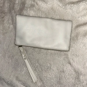 Vit axelväska/plånbok med kedja - Snygg vit axelväska i fakeskinn med guldfärgad kedja och dragkedja. Perfekt för att bära det viktigaste med stil. Väskan har en praktisk rem och stängs med tryckknappar och kedja. Använd bara ett fåtal gånger