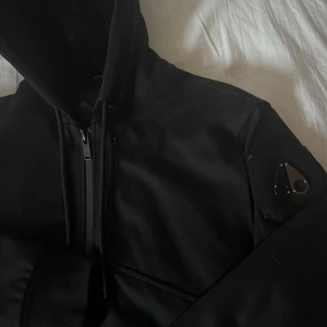 Svart jacka från Moose Knuckles - Säljer en stilren svart jacka från Moose Knuckles med huva och dragkedja. Jackan har ett stort logotypmärke på ryggen och en mindre på ärmen. Den är quiltad inuti för extra värme och har en modern design. Perfekt för kyligare dagar.