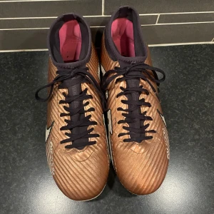 Bronsfärgade fotbollsskor från Nike - Snygga bronsfärgade fotbollsskor från Nike med svart snörning och rosa detaljer på sulan. Skorna har en hög ankelkrage för extra stöd och en iögonfallande design med stora vita logotyper på sidorna.
