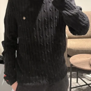 Svart stickad tröja från Morris - Säljer en snygg svart stickad tröja från Morris i storlek S. Den är gjord av merceriserad bomull och har ett diskret emblem på bröstet. Perfekt för höst och vinter med sin stilrena design och långa ärmar. Passar både till vardags och lite finare tillfällen.