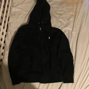 Svart hoodie från Ralph Lauren - Säljer en klassisk svart hoodie från Ralph Lauren med dragkedja och huva. Den har en liten broderad logga på bröstet och är tillverkad i ett mjukt material. Perfekt för en typ alla stilar. Obs fickan är lite sliten och den har några vita fläckar på baksidan men det går bort lätt. Helt ekta ✅✅