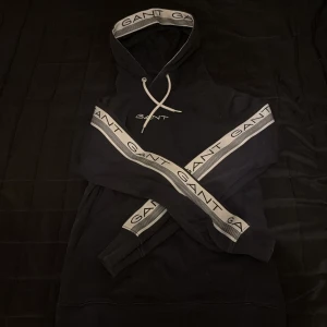 Svart hoodie från GANT - Säljer en svart hoodie från GANT med logotypband på ärmar och huva. Tröjan har en klassisk design med dragsko i huvan och ribbade muddar. Perfekt för en stilren och bekväm look.