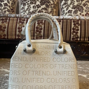 Vit handväska United Colors  - Snygg vit handväska med texten 'UNIFIED COLORS OF TREND' i ett upprepat mönster. Väskan har två handtag och en dragkedja upptill för säker förvaring. Perfekt för att ge en stilren touch till din outfit.