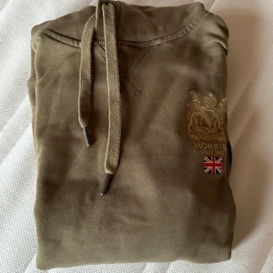 Olivgrön hoodie från Morris - Säljer en olivgrön hoodie från Morris med en broderad logga och brittisk flagga på bröstet. Tröjan har en justerbar huva med snören och en stor ficka framtill. Perfekt för en avslappnad stil.