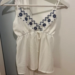 Vit broderad linnetopp - Säljer en söt vit linnetopp med blå broderade blommor längs den v-ringade halsen!