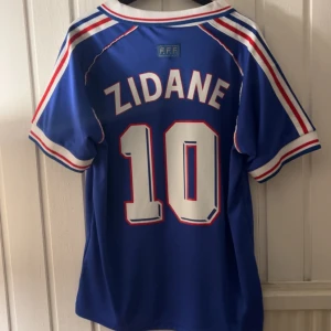 Zidane Fotbollströja - Säljer en blå fotbollströja från Adidas med 'Zidane' och nummer 10 på ryggen. Tröjan har korta ärmar och är dekorerad med vita och röda detaljer. Framsidan har texten 'FIFA World Cup France 98' och det franska landslagets emblem.