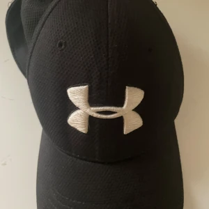 Svart keps från Under Armour - Snygg svart keps från Under Armour med broderad logga framtill. Kepsen har en klassisk design med böjd skärm och justerbar passform. Perfekt för sport och vardag.