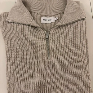 Beige stickad tröja från East West - Säljer en stilren beige stickad tröja från East West med dragkedja vid halsen. Perfekt för kyligare dagar med sin ribbade design och långa ärmar. Ett måste i garderoben för en klassisk look.