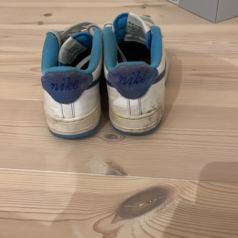 Säljer ett par klassiska Nike Air Force sneakers i vitt med blå detaljer. Skorna har en blå sula och blå insida, samt den ikoniska Nike-loggan i blått på sidan. Perfekta för en sportig look. De är bra skick lite smutsiga kan fixa de om någon vill köpa eller om man vill ta bort de själv men oavsett så är de rätt lätt att få bort. Kengät.